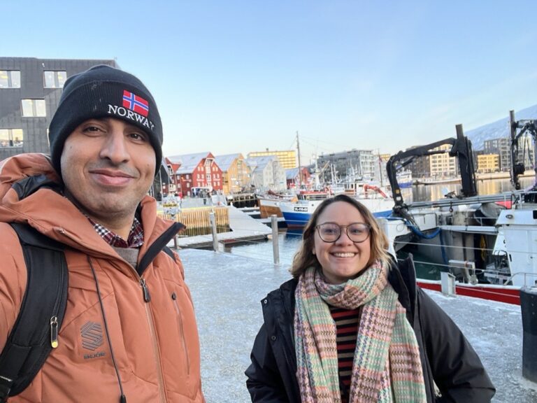 Picture of researchers Morteza and Tangni dressed in warm winter clothes infront of the snowy harbour in Tromsø.